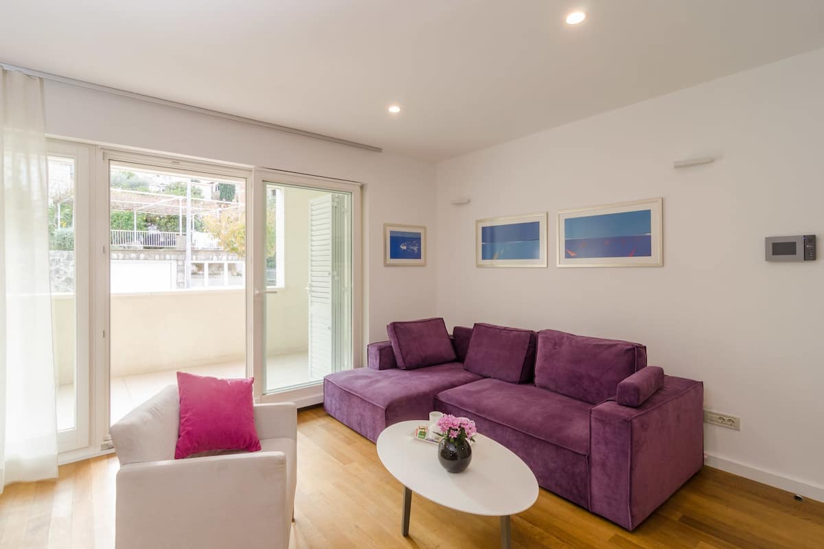 Un salón moderno con un sofá morado, un sillón blanco y una mesita con un florero. Hay dos cuadros enmarcados en la pared y un ventanal corredero que da a un espacio exterior.