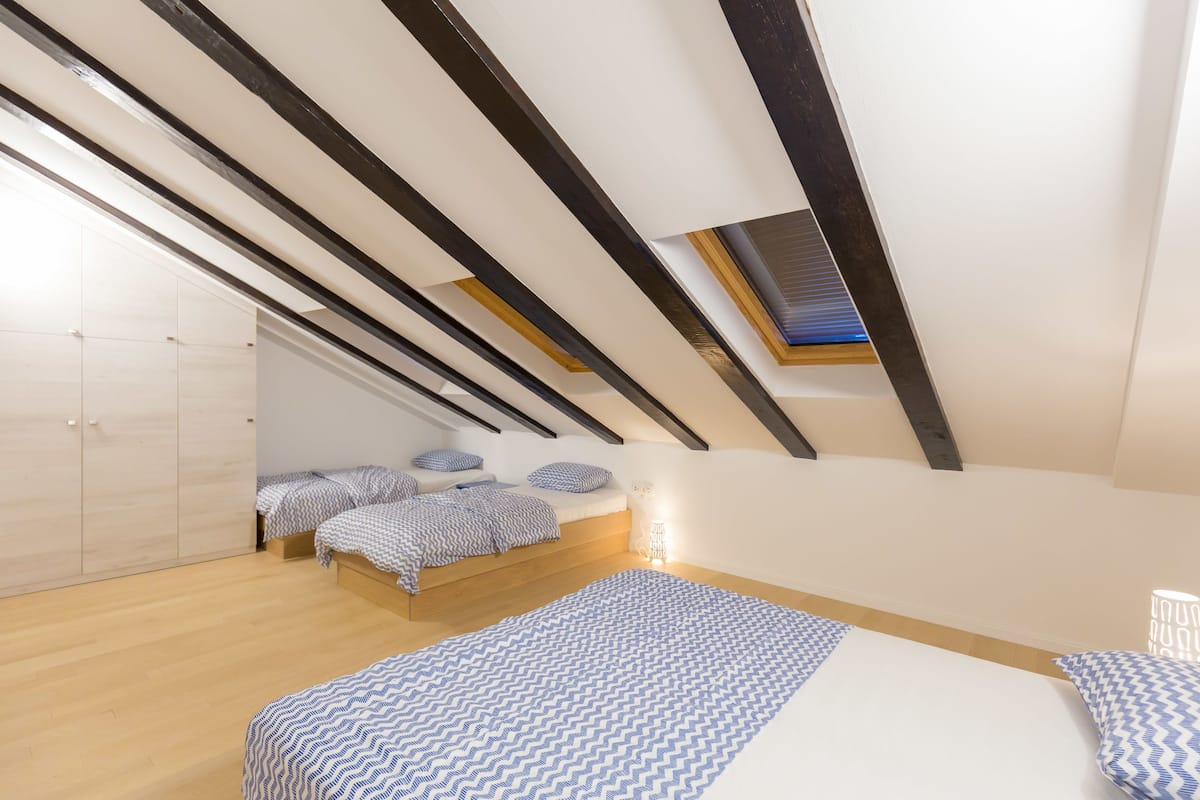 Un dormitorio tipo loft moderno con techo inclinado, vigas de madera, tragaluces y una cama grande con ropa de cama estampada.