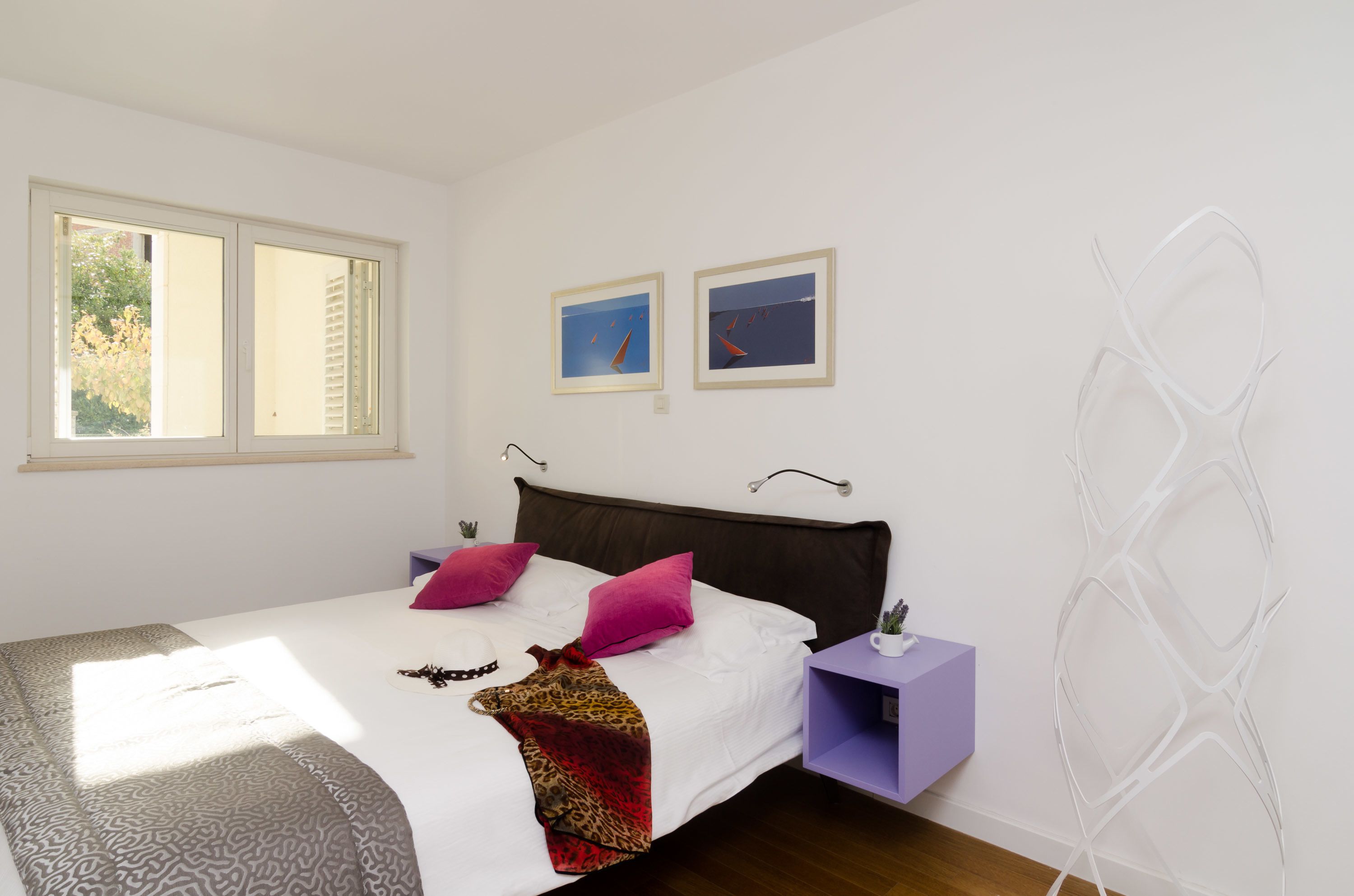 Une chambre à coucher avec un lit, deux oreillers, une table de chevet, une fenêtre et deux tableaux encadrés accrochés au mur.