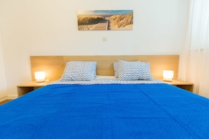 Una cama con manta azul y almohadas estampadas, dos lámparas de noche y un cuadro en la pared que muestra un camino de playa.