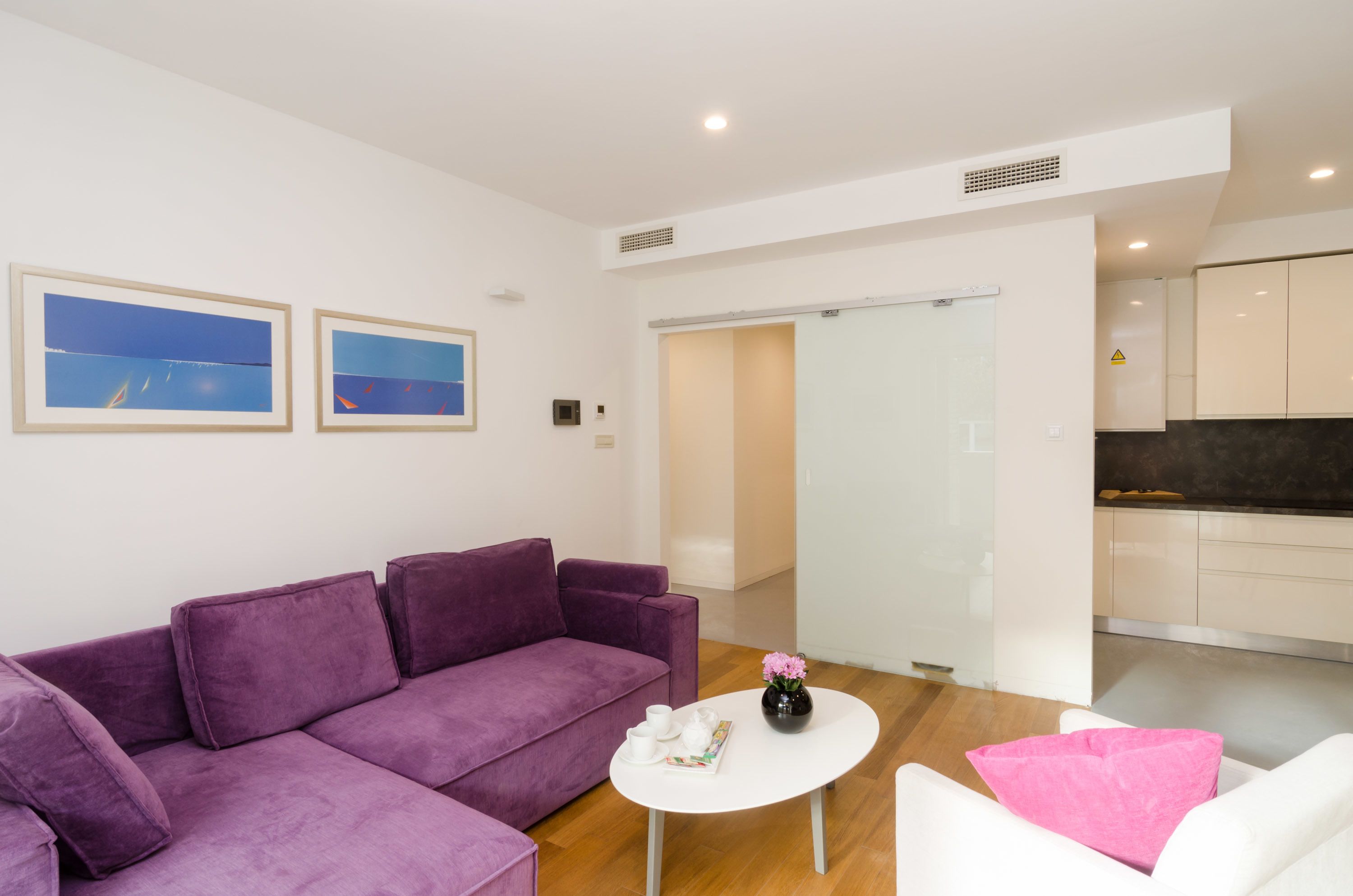 A modern living room with a purple sofa, a white coffee table, and a kitchen area in the background.