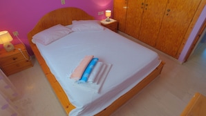 Un lit bien fait, avec des oreillers et des serviettes pliées, une tête de lit en bois, une lampe de chevet et une armoire en bois, dans une pièce aux murs roses.