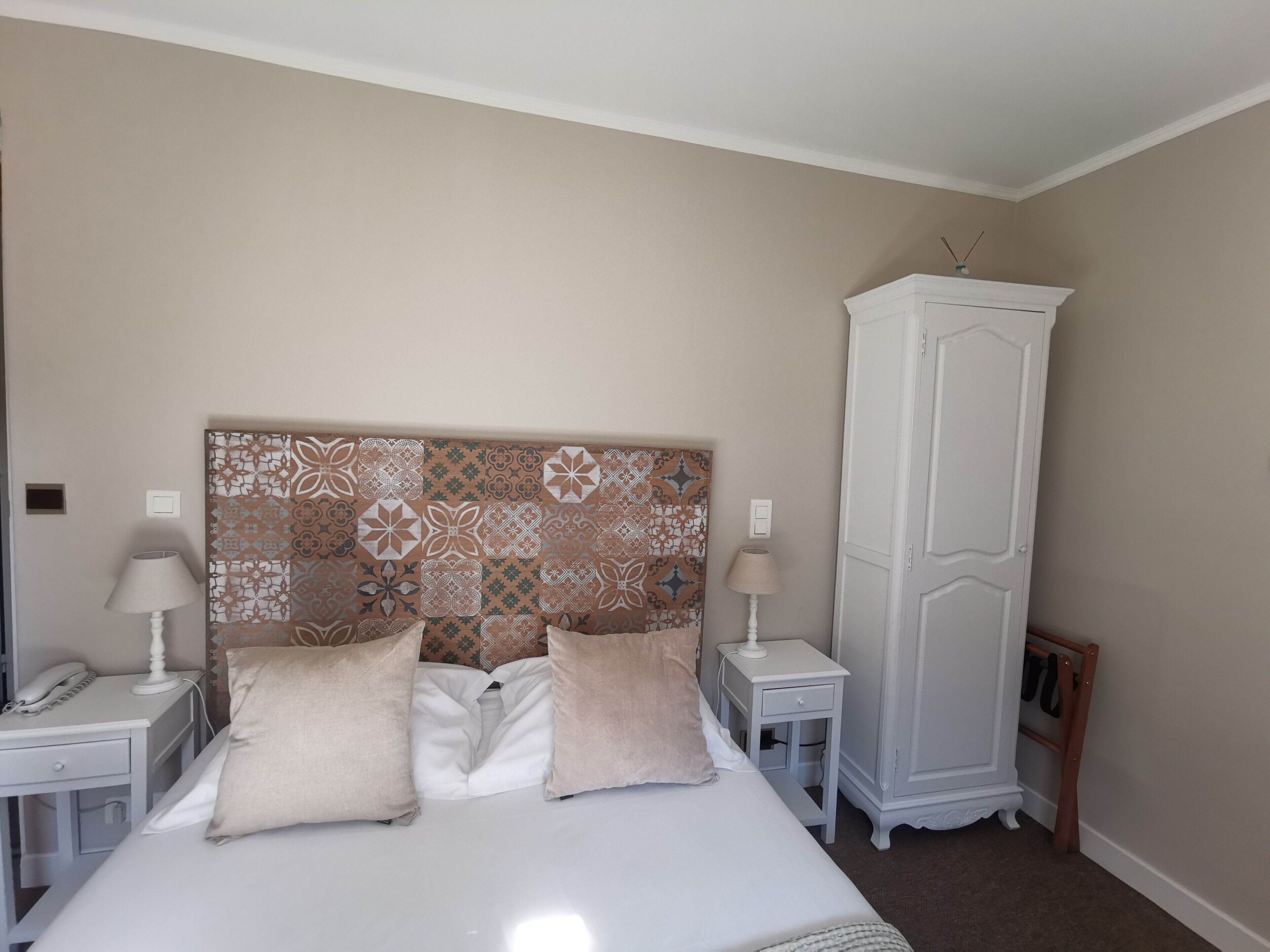 A neatly arranged bedroom with a patterned headboard, two bedside tables with lamps, a wardrobe, and a bed with two pillows.