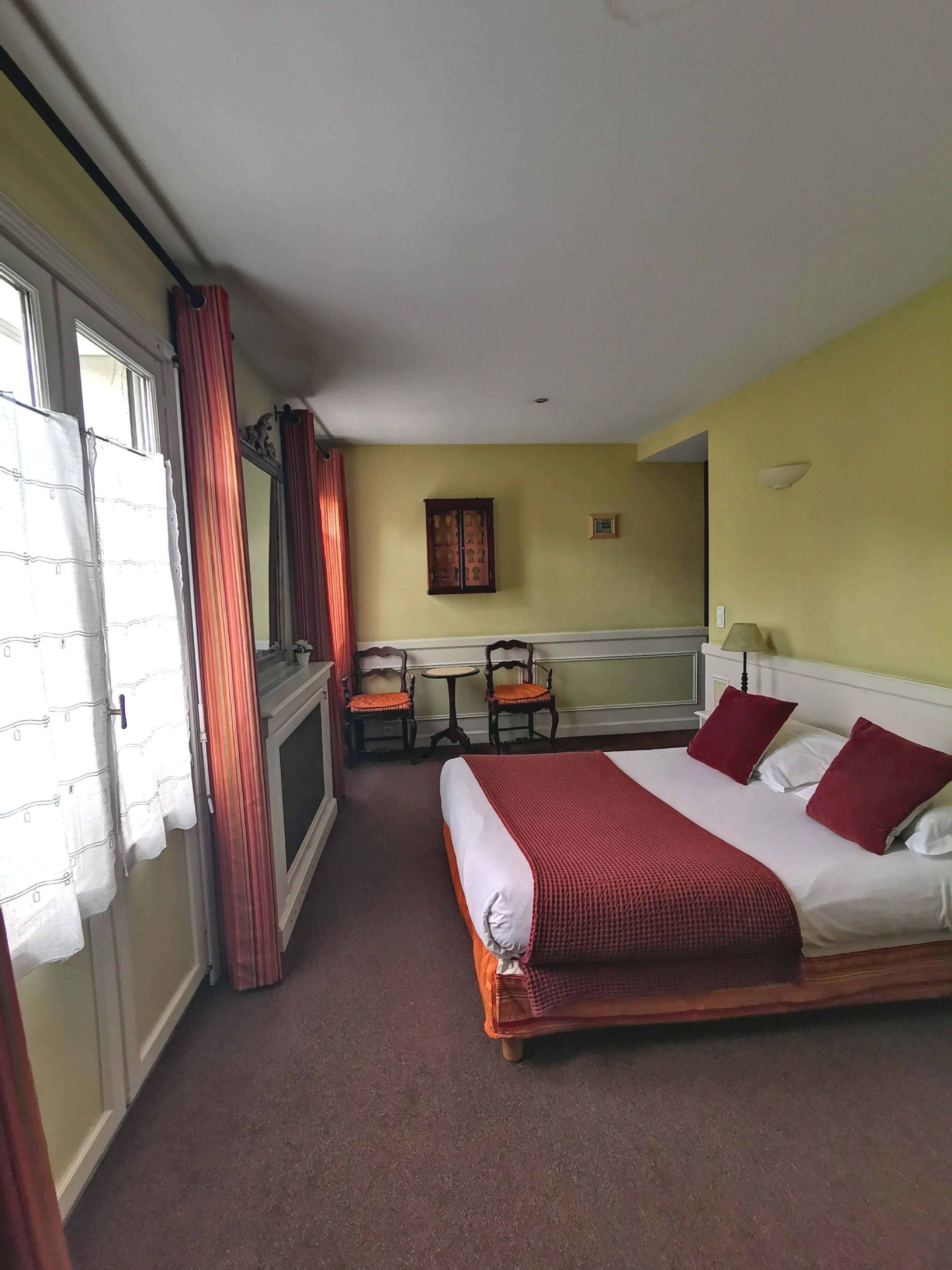 Quarto de hotel com uma cama, duas cadeiras, uma mesinha, um armário e uma janela com cortinas.