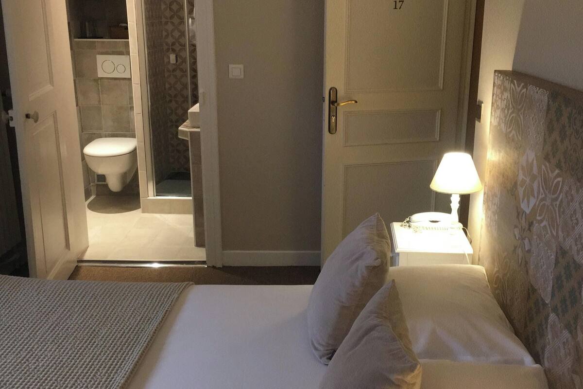 A hotel room with a bed, a nightstand with a lamp, a bathroom with a toilet and shower, and a door with a number.