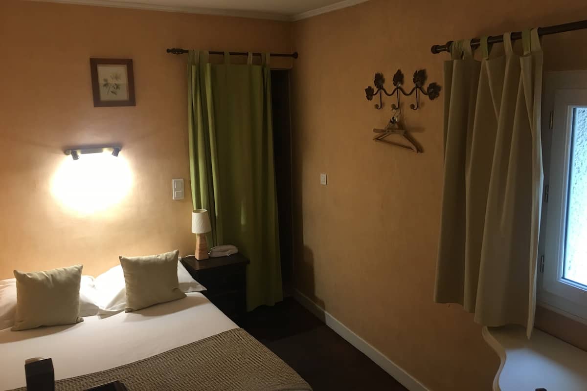 A hotel room with a bed, a nightstand, a lamp, a wall-mounted light, a window with curtains, and a wall decoration.