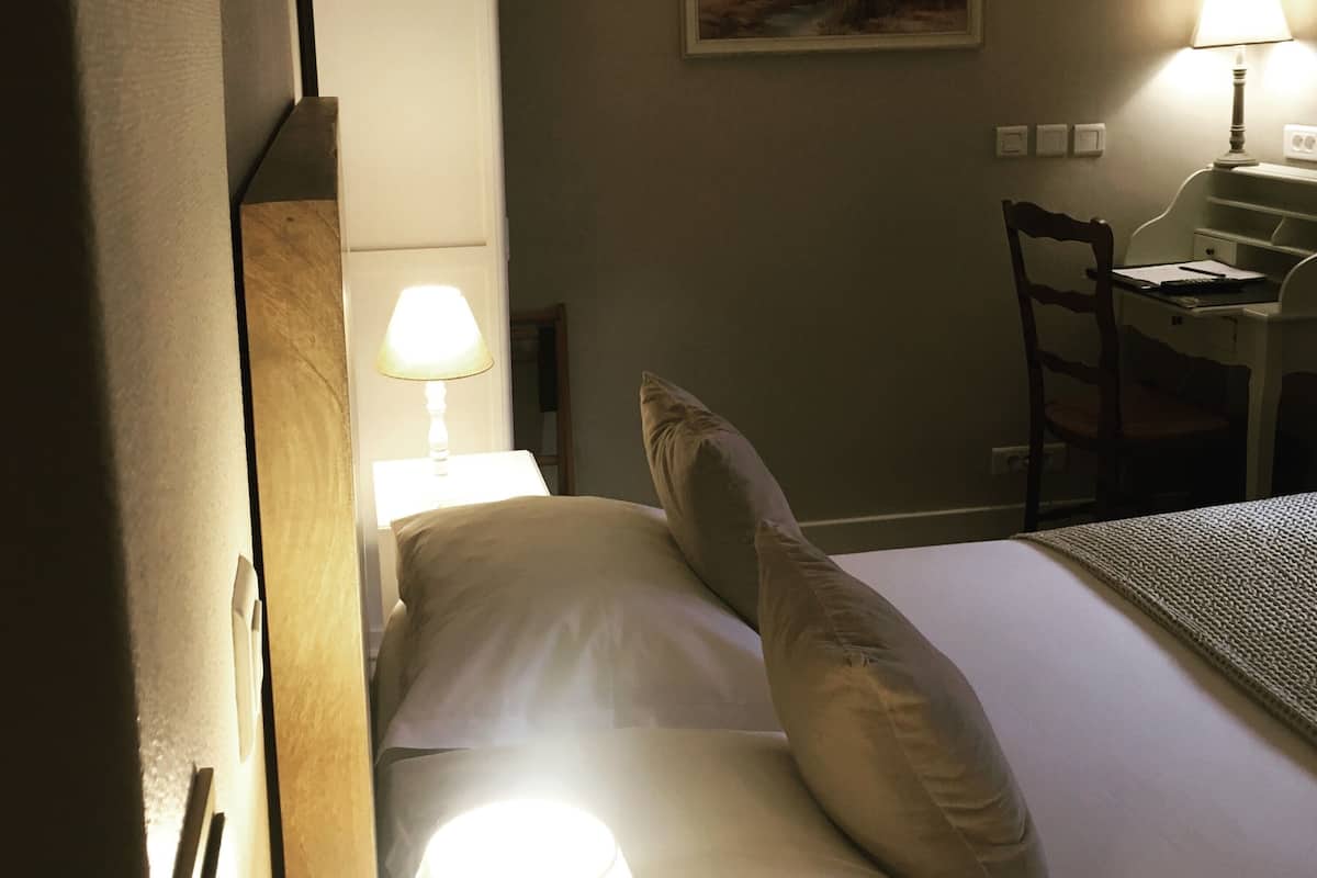 A hotel room with a bed, a desk with a chair, a painting on the wall, and a lamp on the desk.