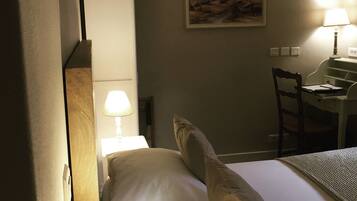 Quarto de hotel com uma cama, uma escrivaninha com cadeira, um quadro na parede e uma luminária sobre a escrivaninha.