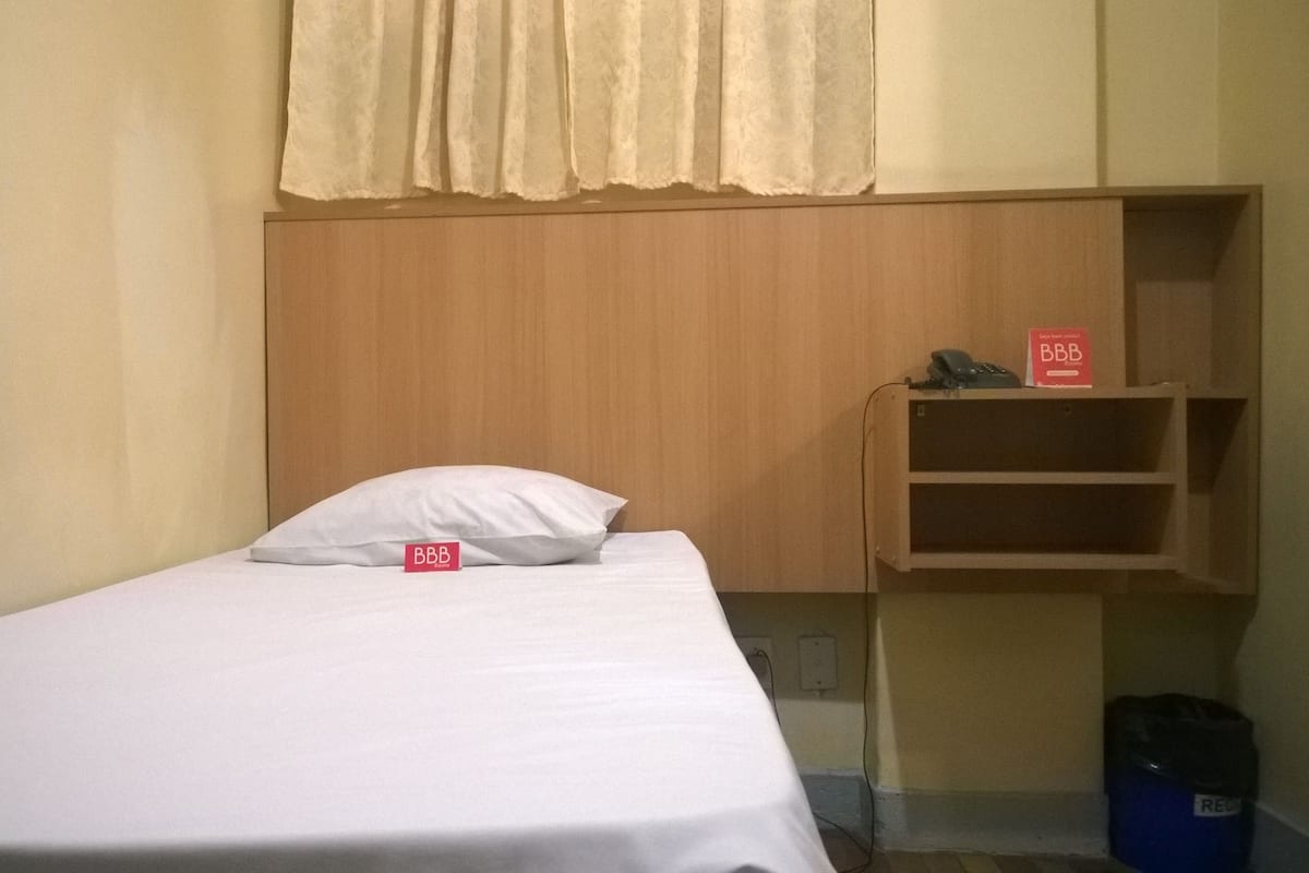 Un lit simple avec un matelas et un oreiller blancs, une tête de lit en bois, une petite étagère avec un téléphone et un panneau, et une poubelle bleue.