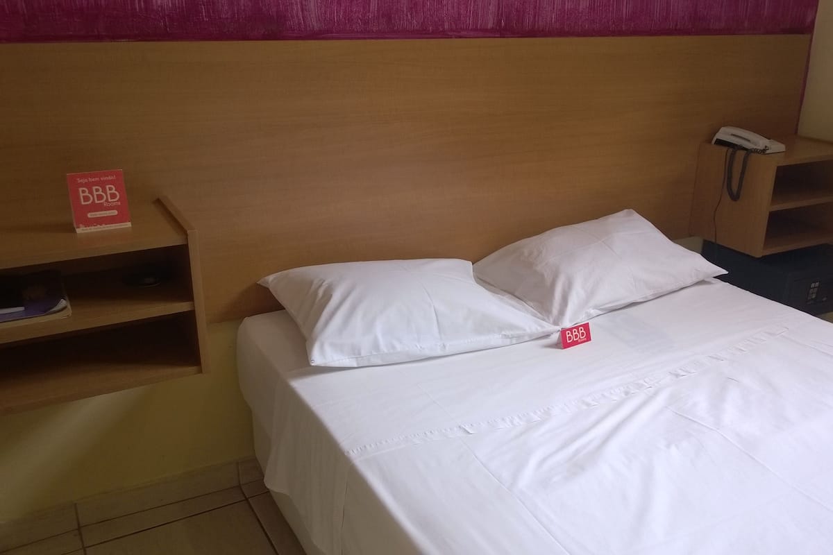 Une chambre d’hôtel avec une tête de lit en bois, un lit blanc avec deux oreillers, une table de chevet avec un téléphone et une étagère sur laquelle est posé un panneau indiquant « BBB ».