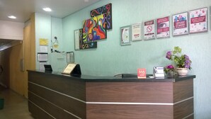 Ein Empfangsschalter mit Holzoptik, darüber ein farbenfrohes abstraktes Gemälde und mehrere Informationsplakate an der Wand.