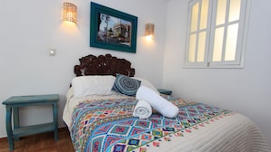 Uma cama bem arrumada com um edredom colorido, duas mesas de cabeceira, um quadro na parede e uma janela com molduras brancas.