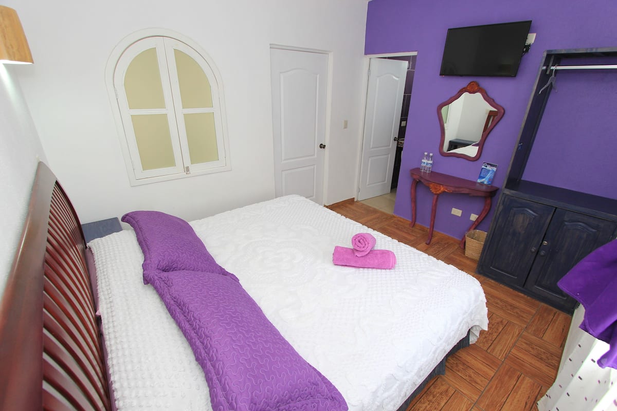 A bedroom with a bed, a wooden headboard, a window, a mirror, a TV, and a small table with a basket.