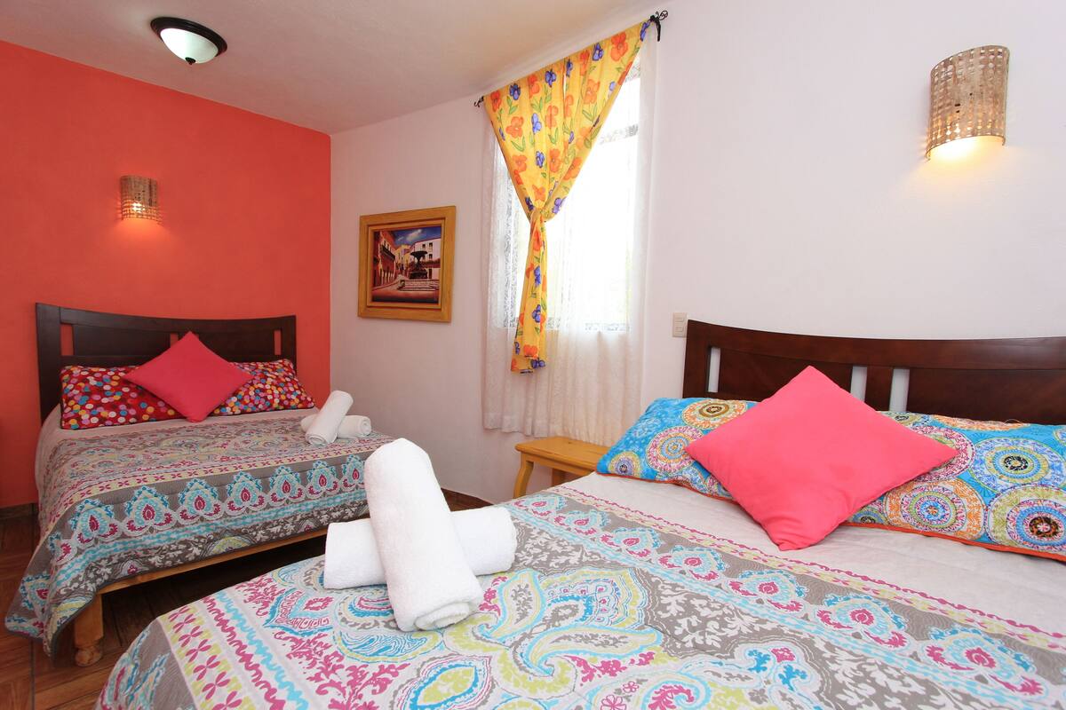 A hotel room with two beds, each with patterned bedding and pillows, a small wooden table, and a wall-mounted light fixture.