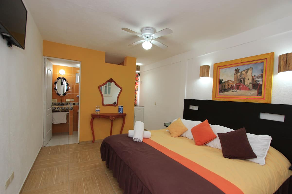 A hotel room with a bed, a ceiling fan, a painting, and a bathroom visible through an open door.