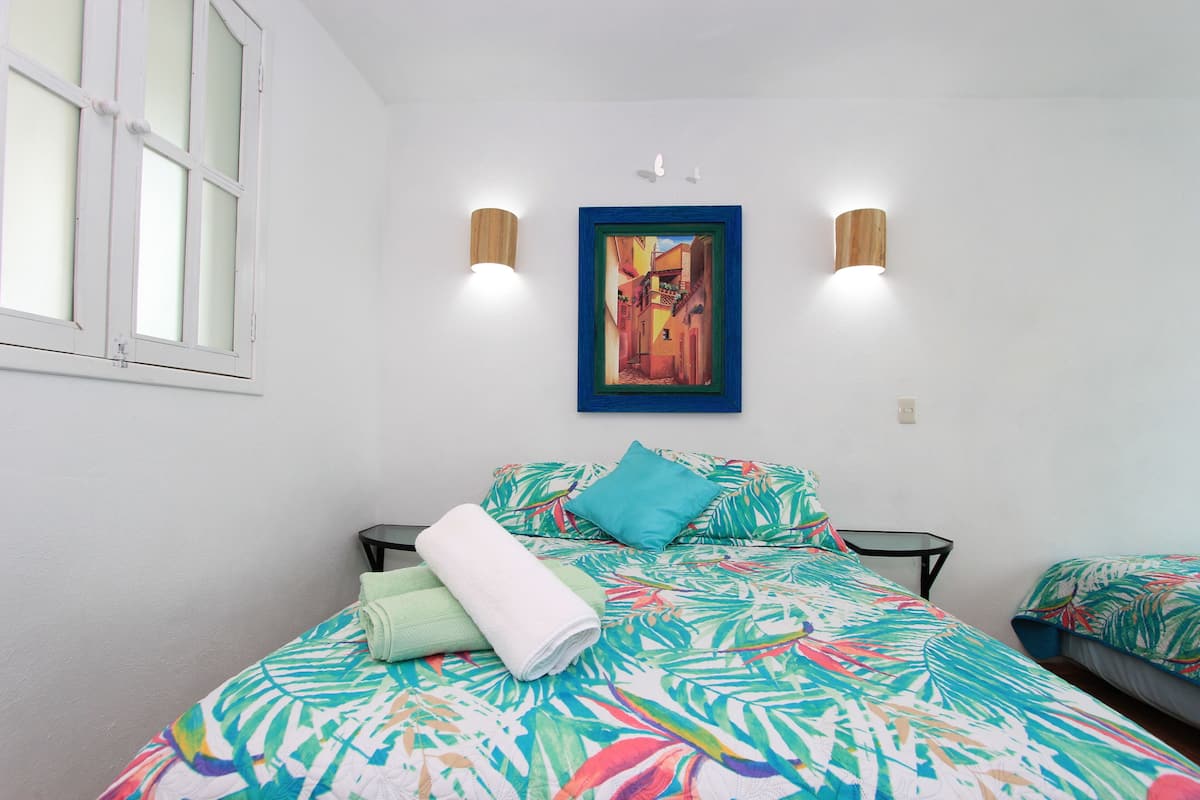 A bedroom with a bed, two wall-mounted lights, a framed painting, and a window.