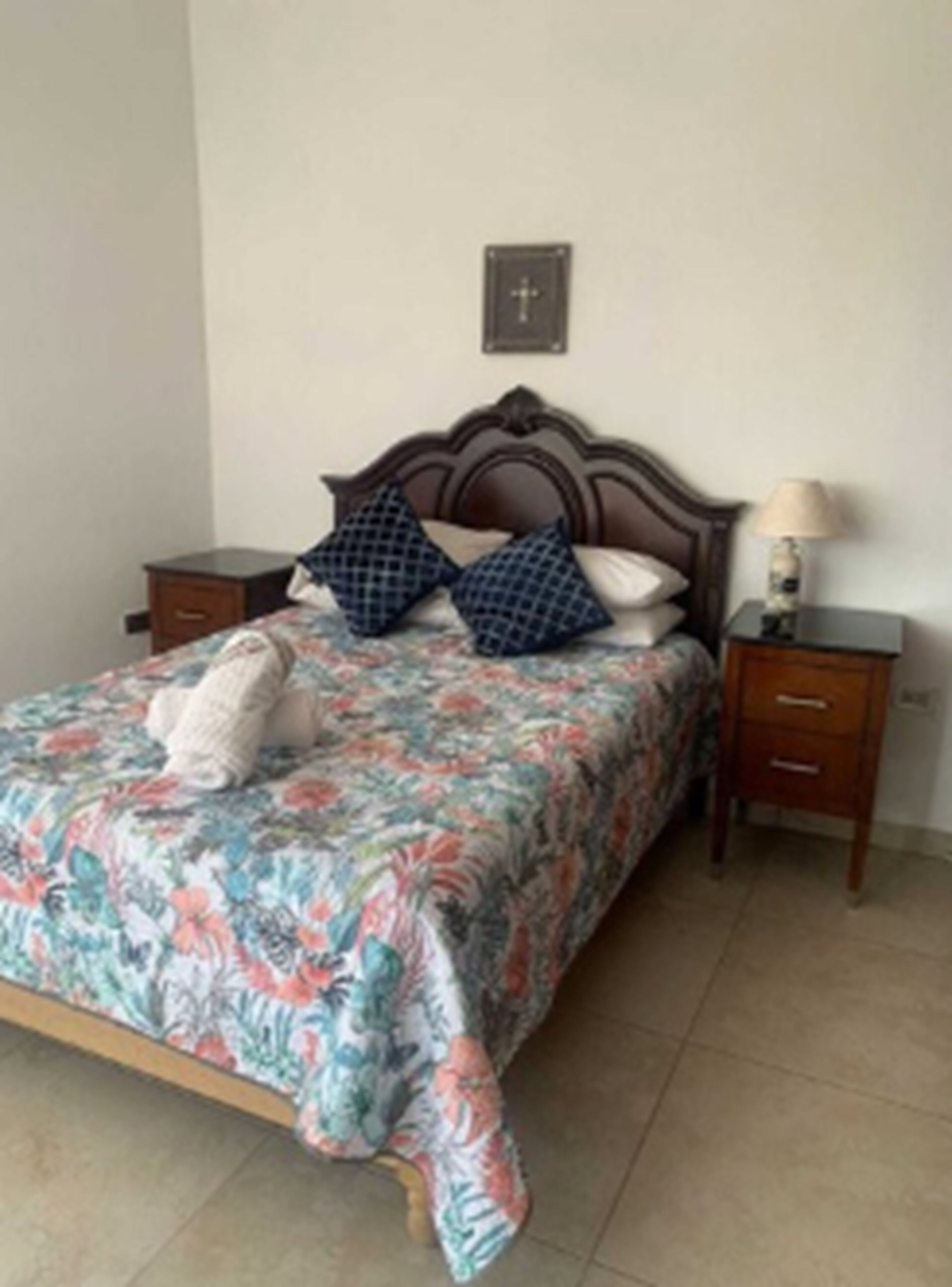 A bedroom with a floral bedspread, two pillows, a wooden headboard, a bedside table with a lamp, and a small wooden nightstand.