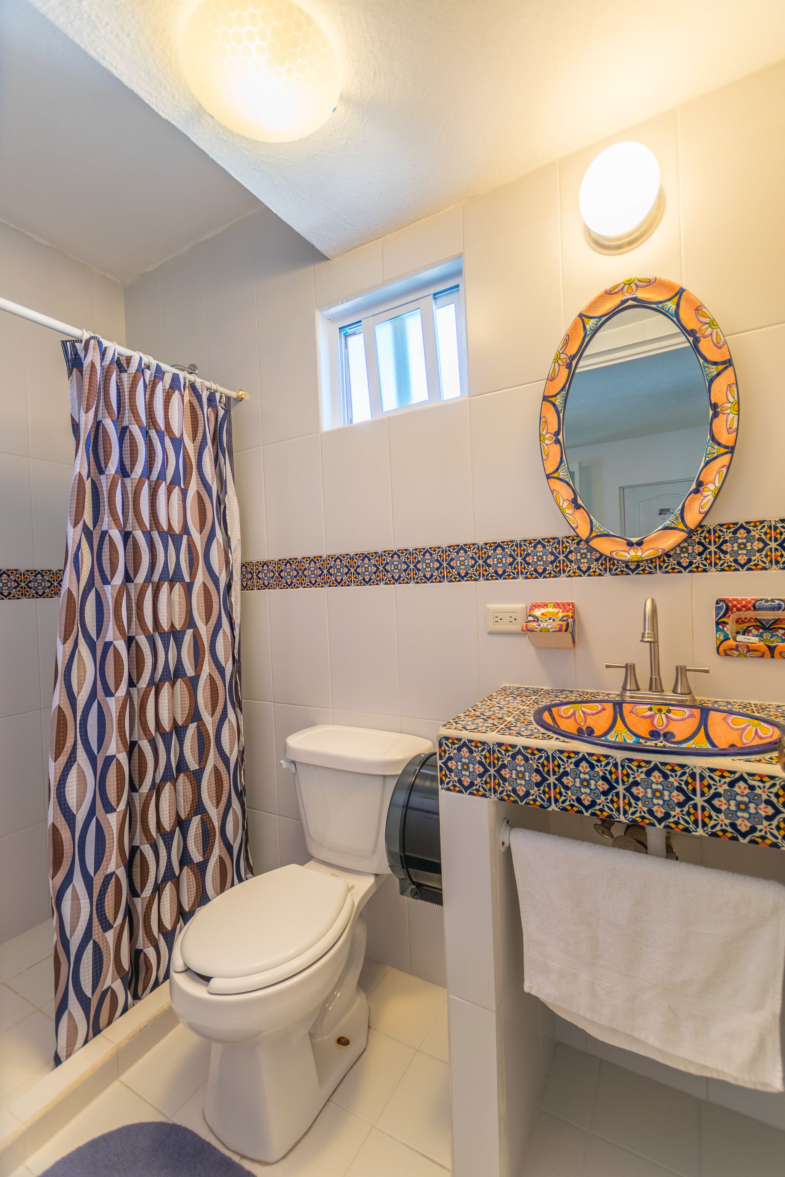 A bathroom with a patterned shower curtain, a toilet, a sink with a decorative tile backsplash, and a mirror with an ornate frame.