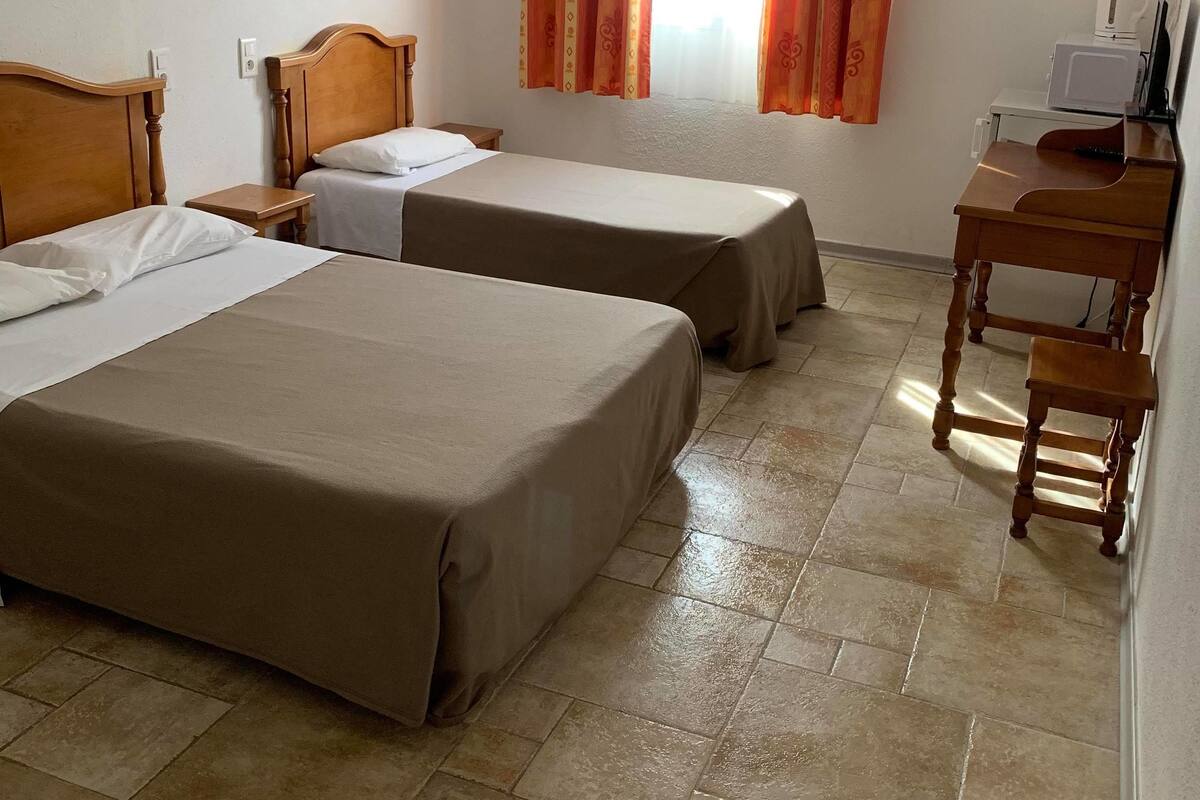 A hotel room with two single beds, a wooden nightstand, a small table, an air conditioning unit, and a window with curtains.