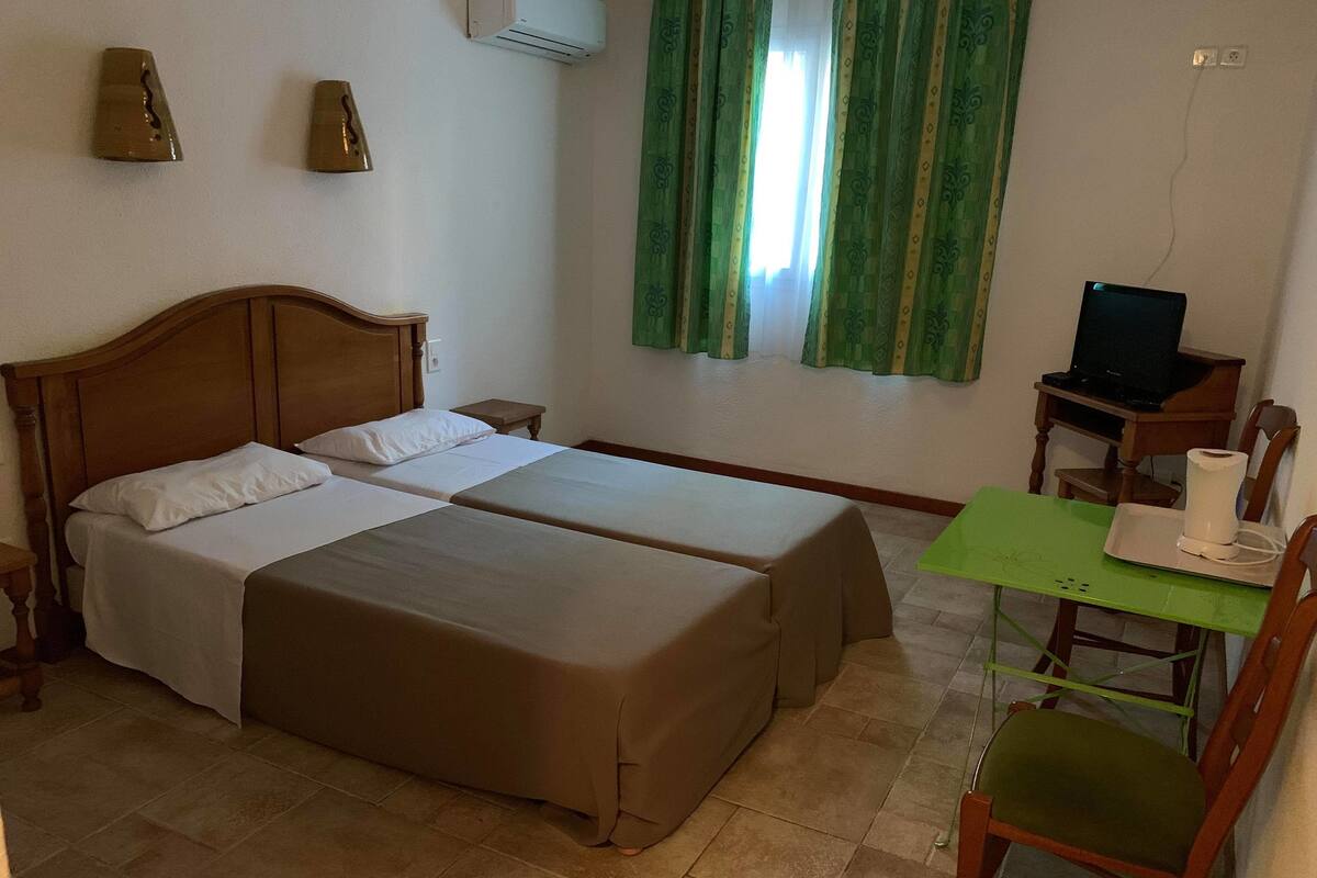 A double bed with a wooden headboard, a small bedside table, a green chair, a desk with a computer, and a TV.