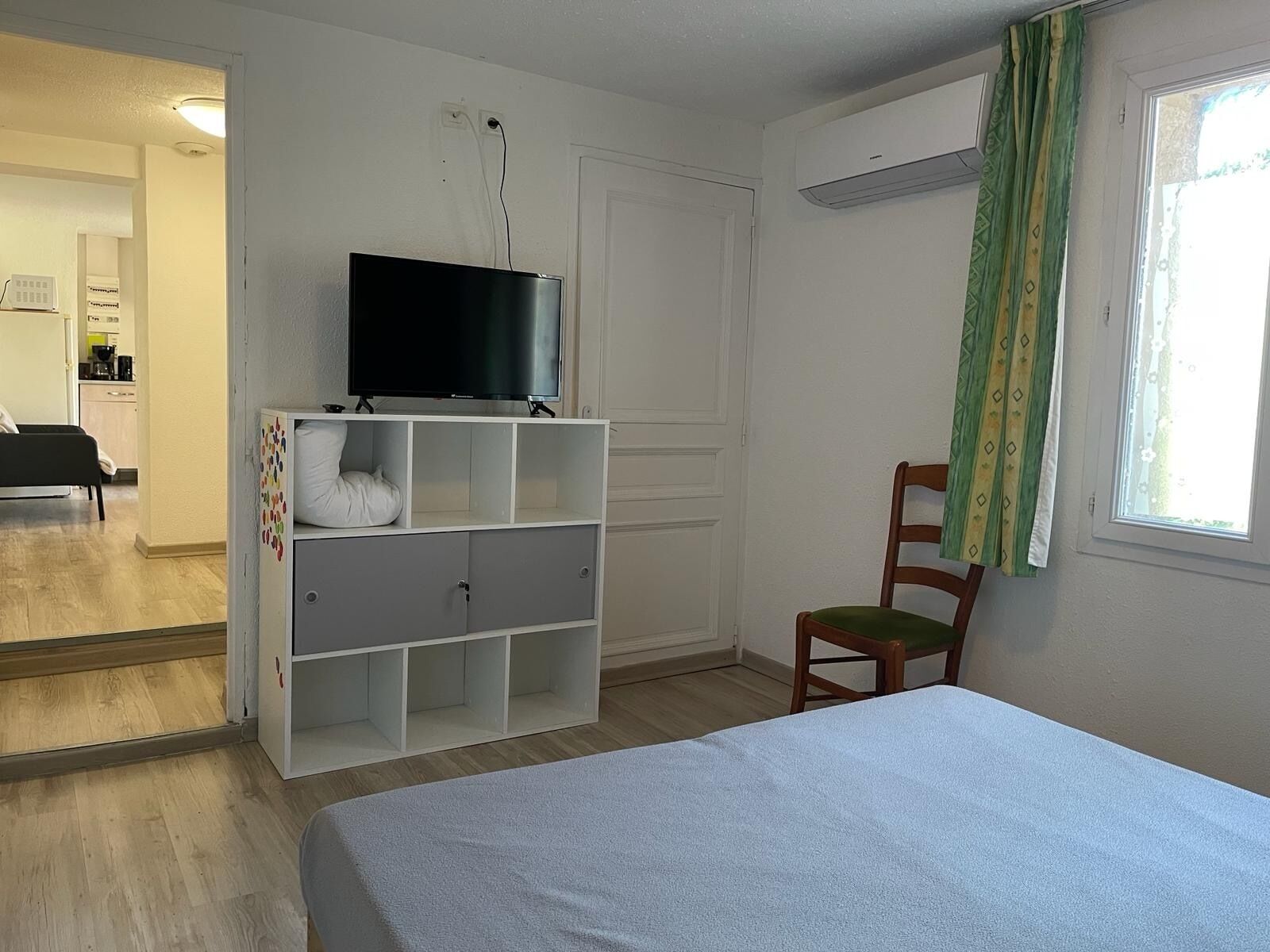 A bedroom with a bed, a TV on a white cabinet, a chair, and a window with curtains.