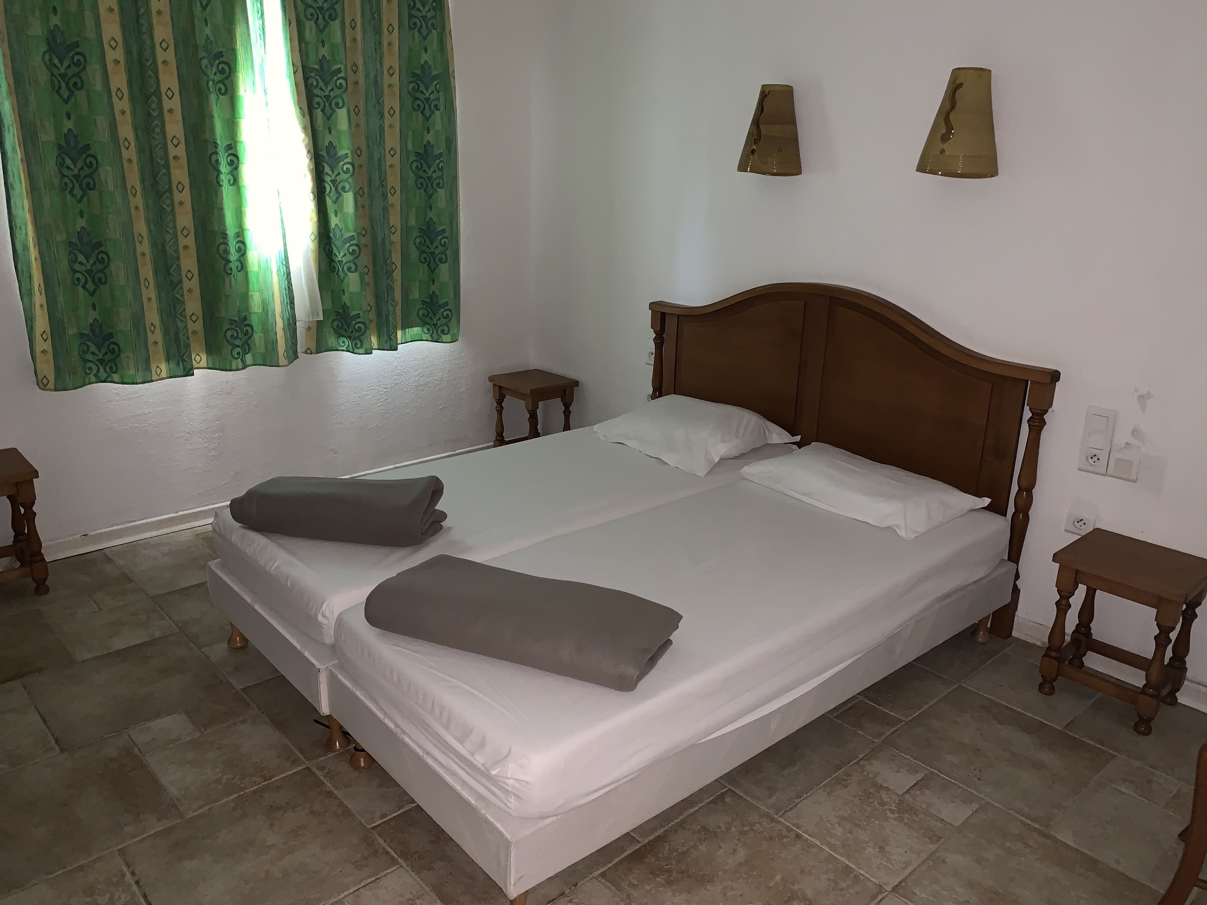 A neatly made bed with pillows and a blanket, a wooden headboard, a small bedside table, and a window with green curtains.