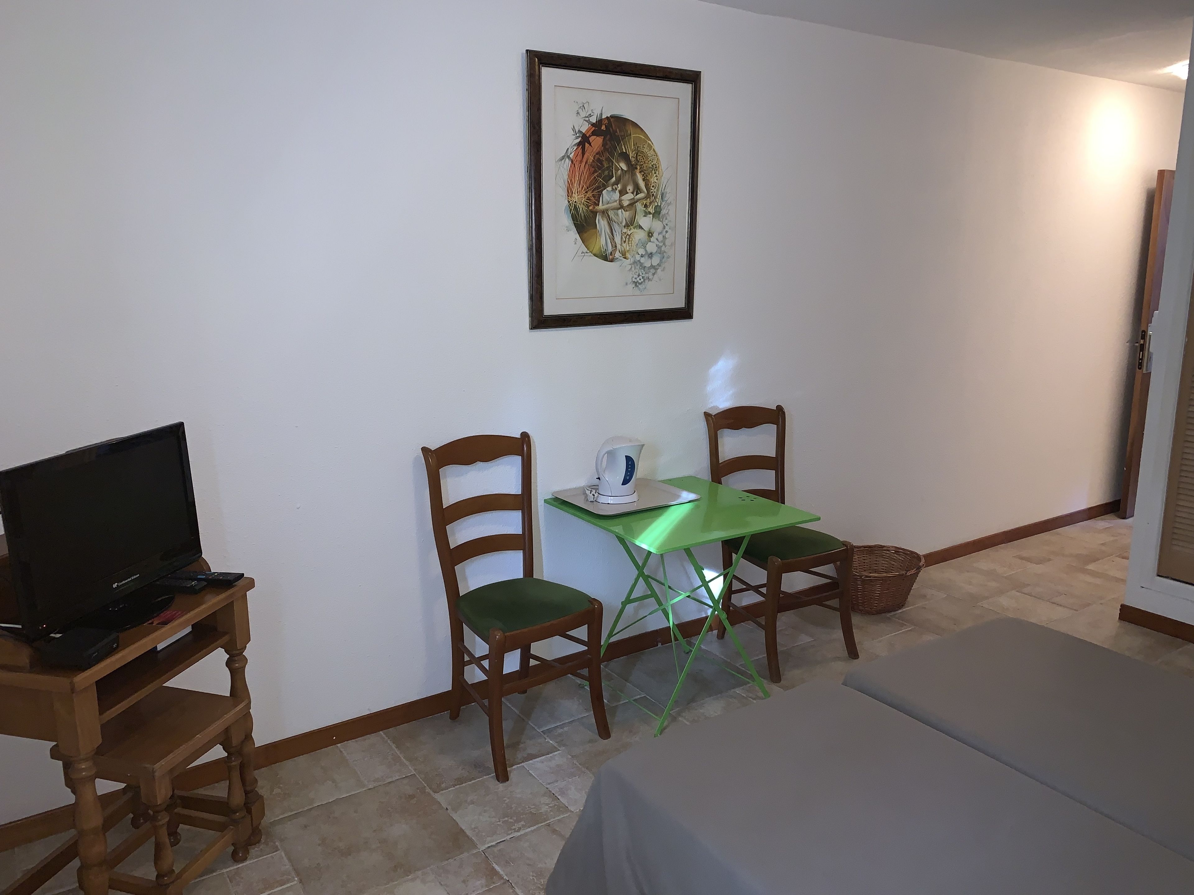 Una habitación pequeña con cama, televisión, dos sillas de madera, una mesita con tetera y un cuadro enmarcado en la pared.