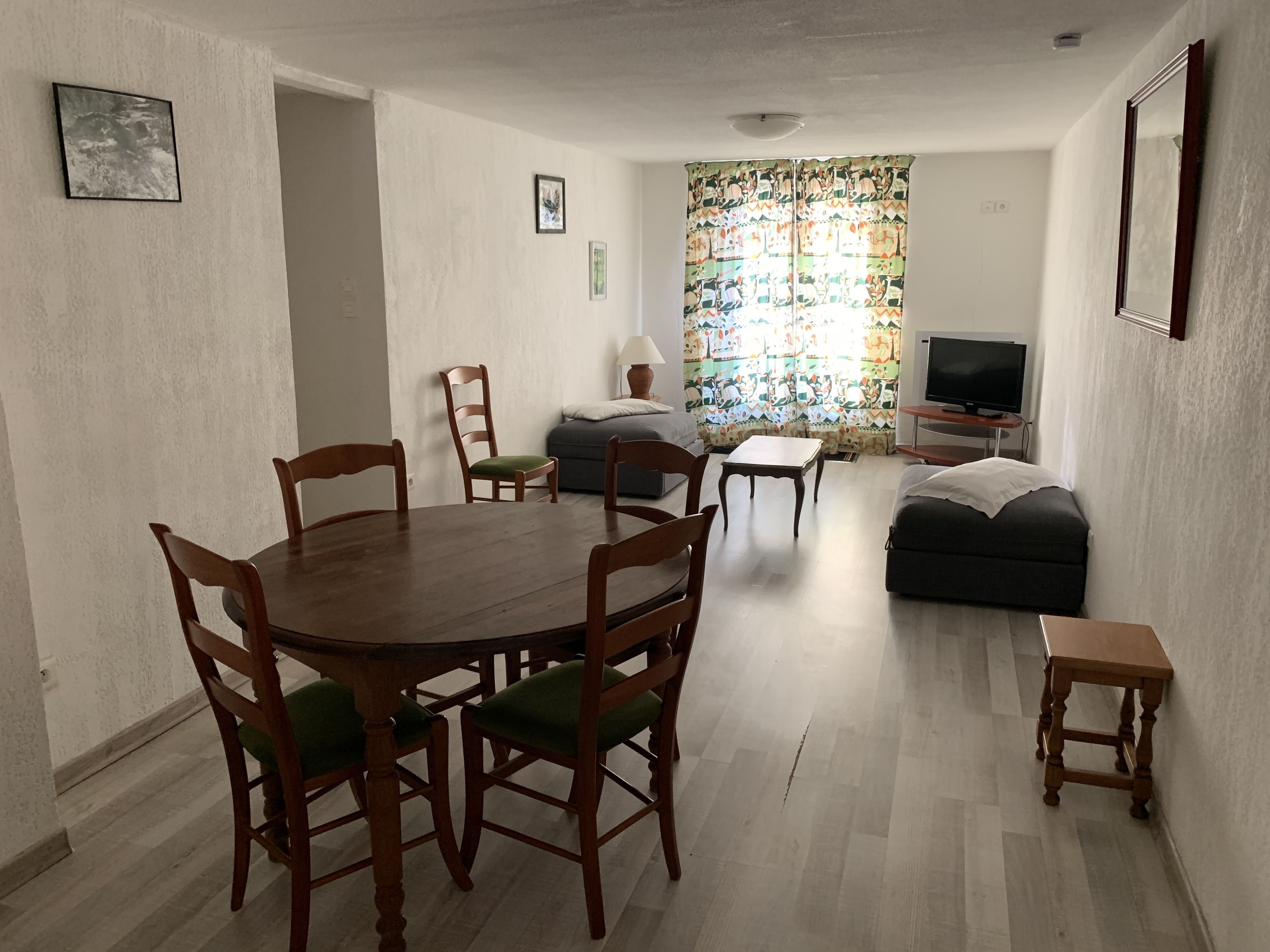 Ein Wohnzimmer mit Esstisch, Stühlen, einer Couch, einem Fernseher und einem Fenster mit Vorhängen.