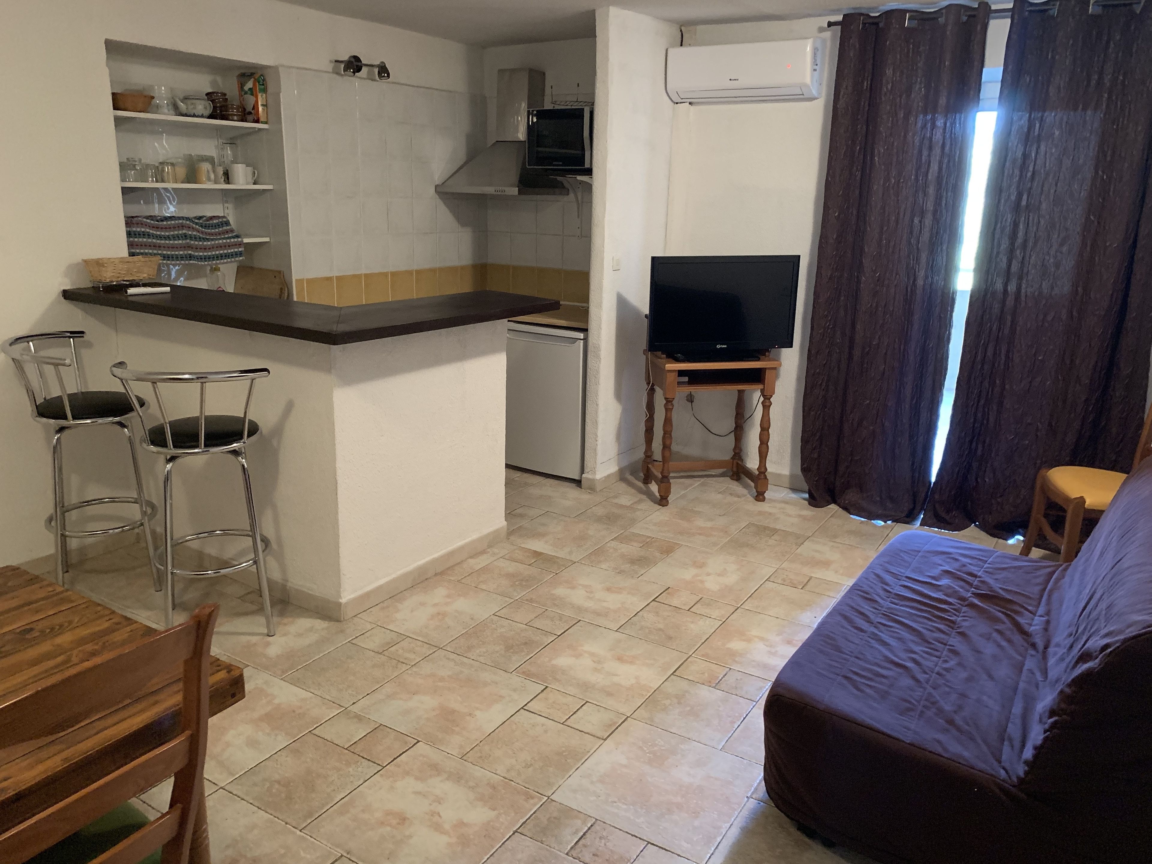 A compact living space with a kitchenette, a dining area with bar stools, a television on a wooden stand, and a bed.