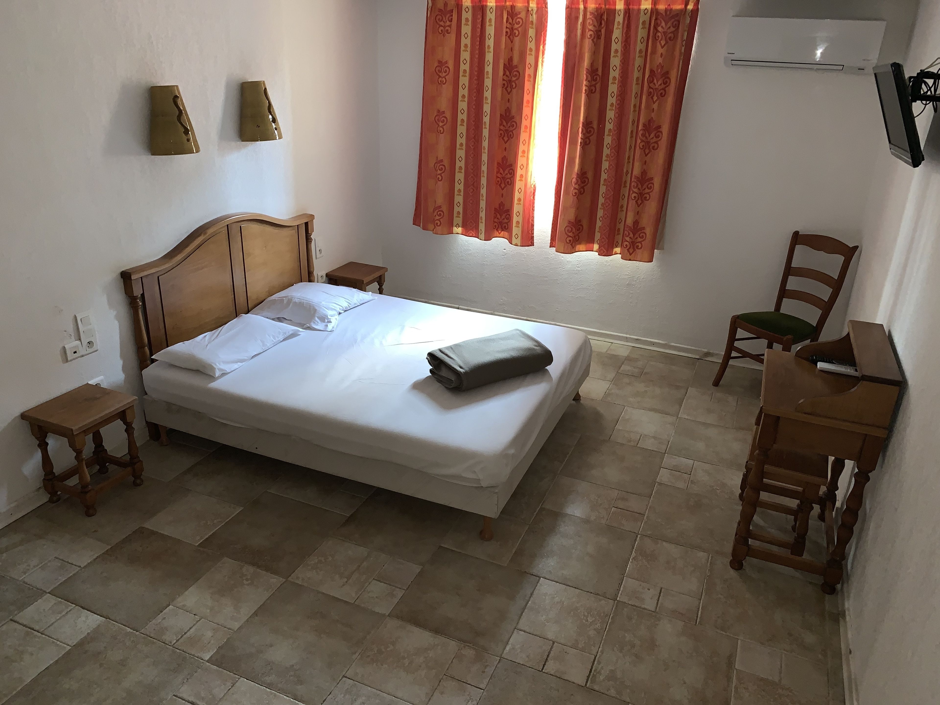 A bedroom with a wooden bed, a small bedside table, a chair, a desk, and a window with curtains.