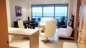 Un moderno salón con comedor, un sofá blanco, una mesa de centro y vistas al océano a través de amplios ventanales.