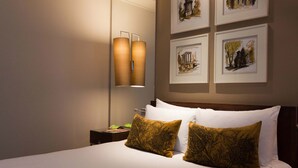 Una habitación de hotel con una cama grande, dos almohadones decorativos, una mesita de noche, una lámpara de pared y una obra de arte enmarcada sobre la cama.