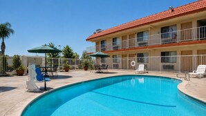 A swimming pool with lounge chairs and umbrellas, surrounded by a fenced patio area with a building in the background. - Vagabond Inn Bakersfield North (Bakersfield)