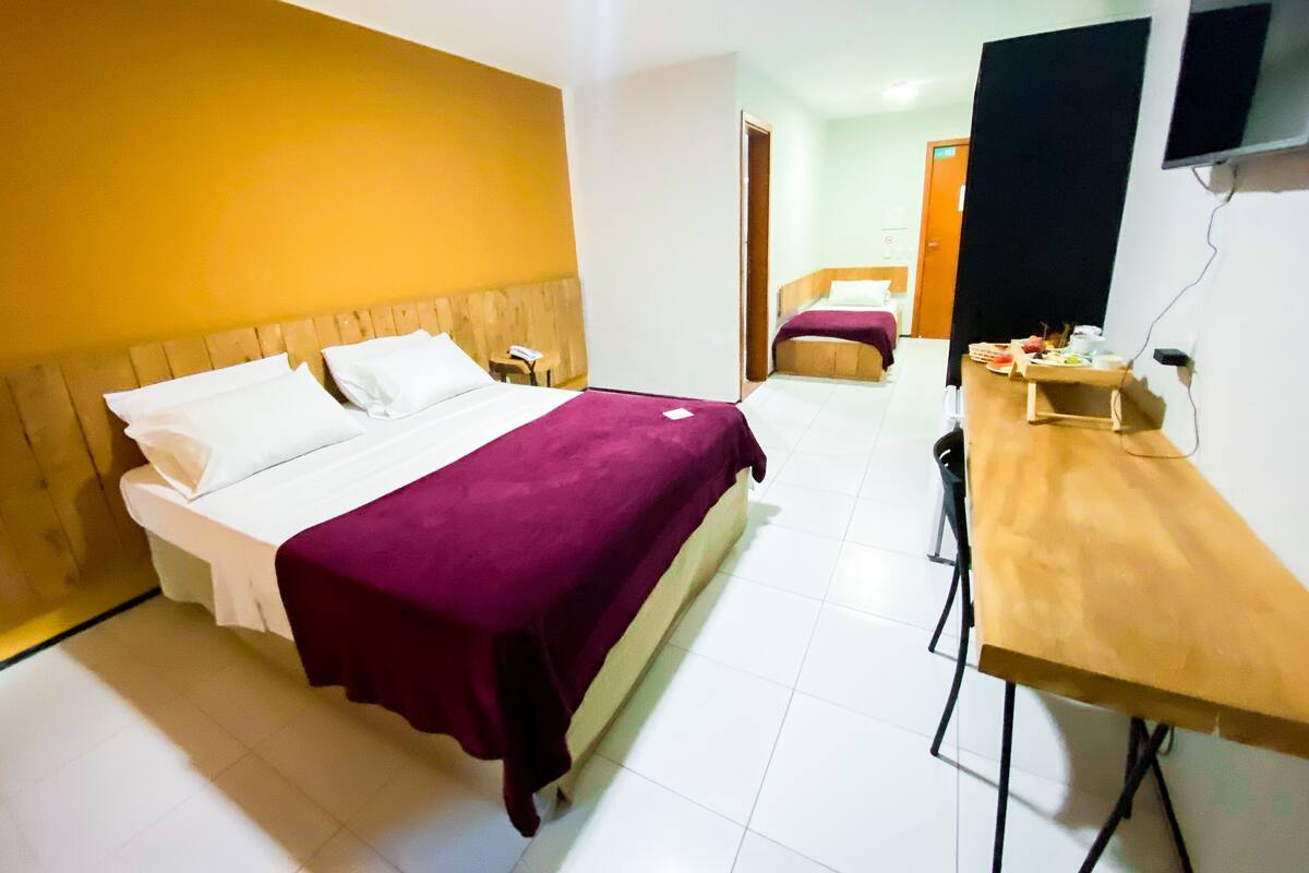 A hotel room with a bed, a desk, a chair, a TV, and a small table with food items.
