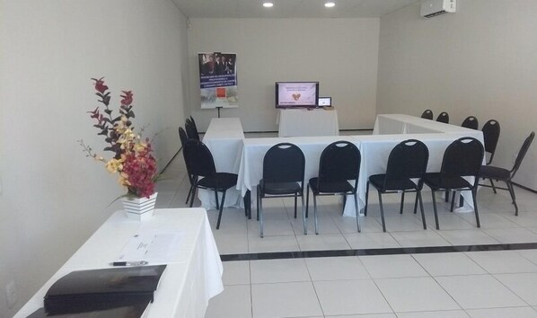Un salón de conferencias con una larga mesa, sillas negras y un mantel blanco. Hay un cartel en la pared y un monitor de computadora.