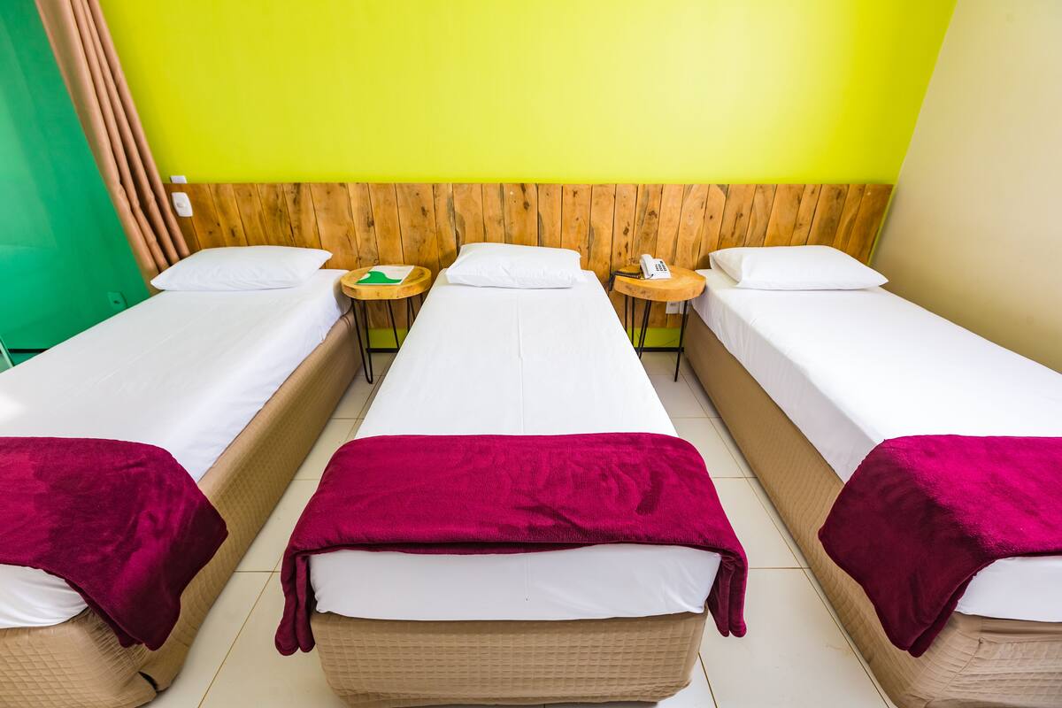 A room with three single beds, each with a wooden headboard and a white mattress. There are two small round tables with a phone and a bottle on each. The walls are painted green and yellow.