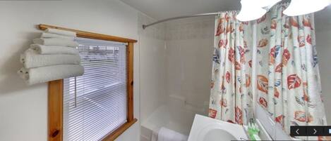 Un baño con ducha, una ventana con persianas y una cortina con estampado de peces.