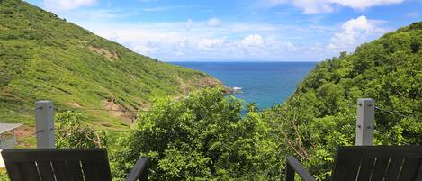 A balcony with two wooden chairs overlooking a coastal landscape with lush greenery and a view of the ocean.