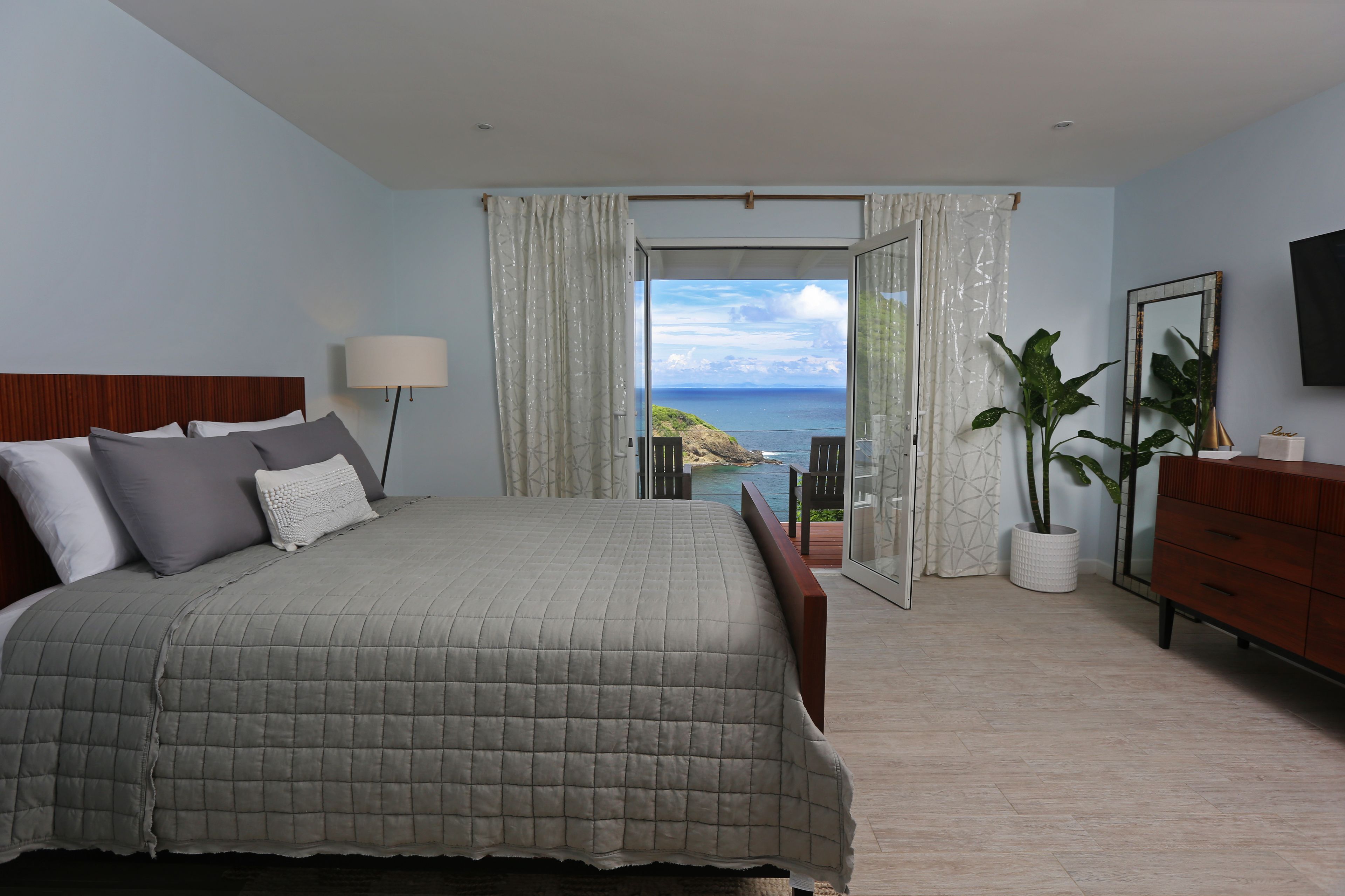 Un dormitorio con una cama grande, una mesita de noche, una lámpara, un aparador y vistas al mar a través de una puerta de cristal.