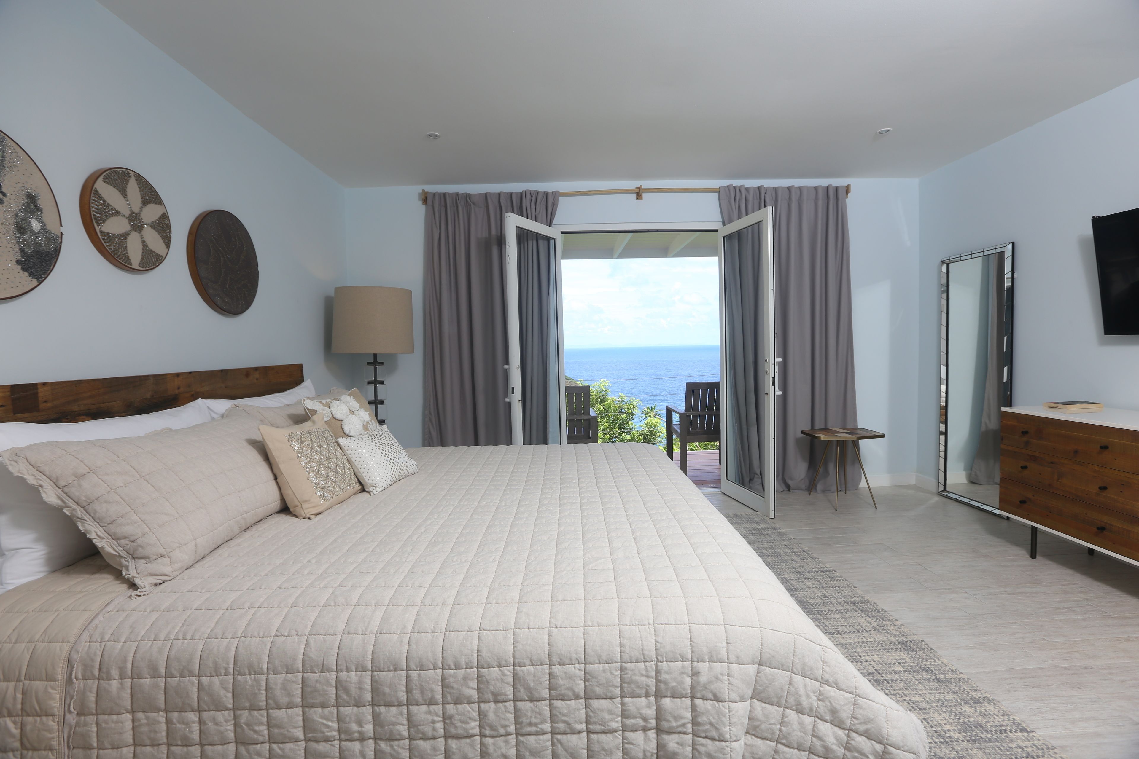 Une chambre à coucher avec un grand lit, une tête de lit en bois, une table de chevet avec une lampe, une commode et une vue sur la mer par une porte ouverte.