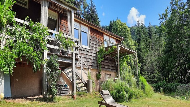 A rustic wooden house with a balcony, a wooden deck, and a garden area with a lounge chair.