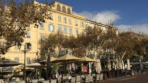 Ein Straßencafé mit Korbtischen und -stühlen, umgeben von Bäumen und einem gelben Gebäude mit Rundbogenfenstern.