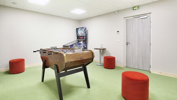 Ein Zimmer mit einem Kicker, zwei roten Hocker, einem an der Wand montierten Fernseher und einer Tür mit einem Notausgangsschild.