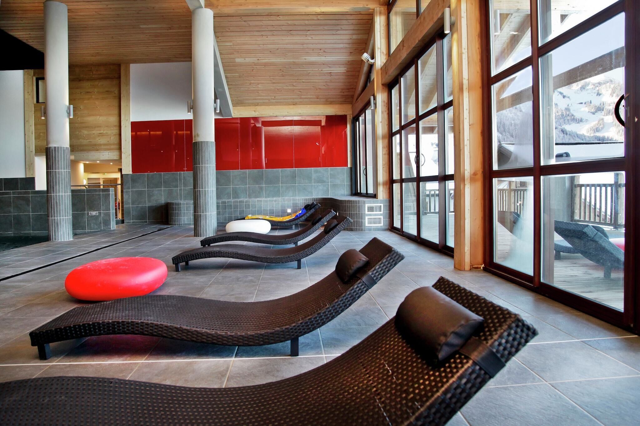 Un área de spa con sillones reclinables, un reposapiés rojo y grandes ventanales con vistas a un paisaje nevado.