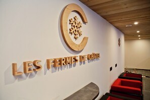Ein Empfangsbereich mit einem hölzernen Schild, das 'LES FERMES DE CHATEAU' zeigt, und einem Logo darüber. Es gibt rote Sitzbereiche und eine Holzbalkendecke.