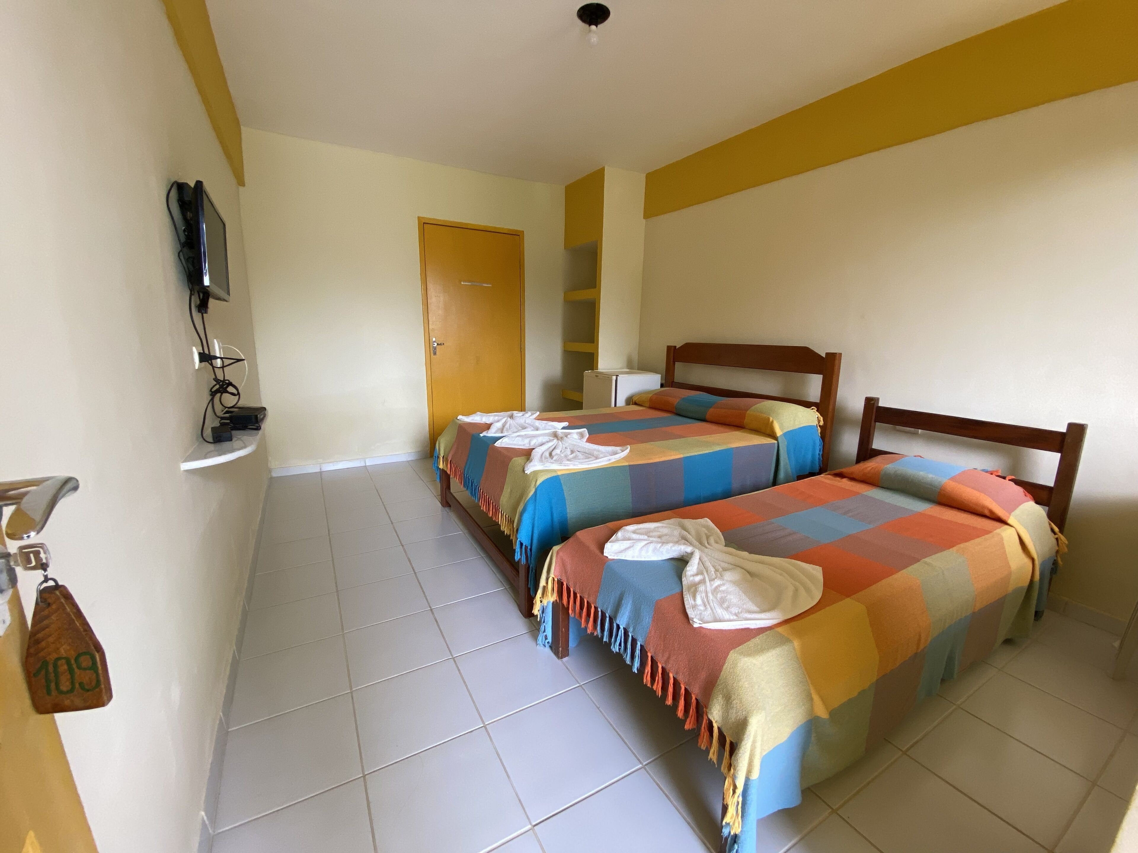 Habitación de hotel con dos camas, un televisor fijado a la pared, un refrigerador pequeño y una puerta que da a otra habitación.
