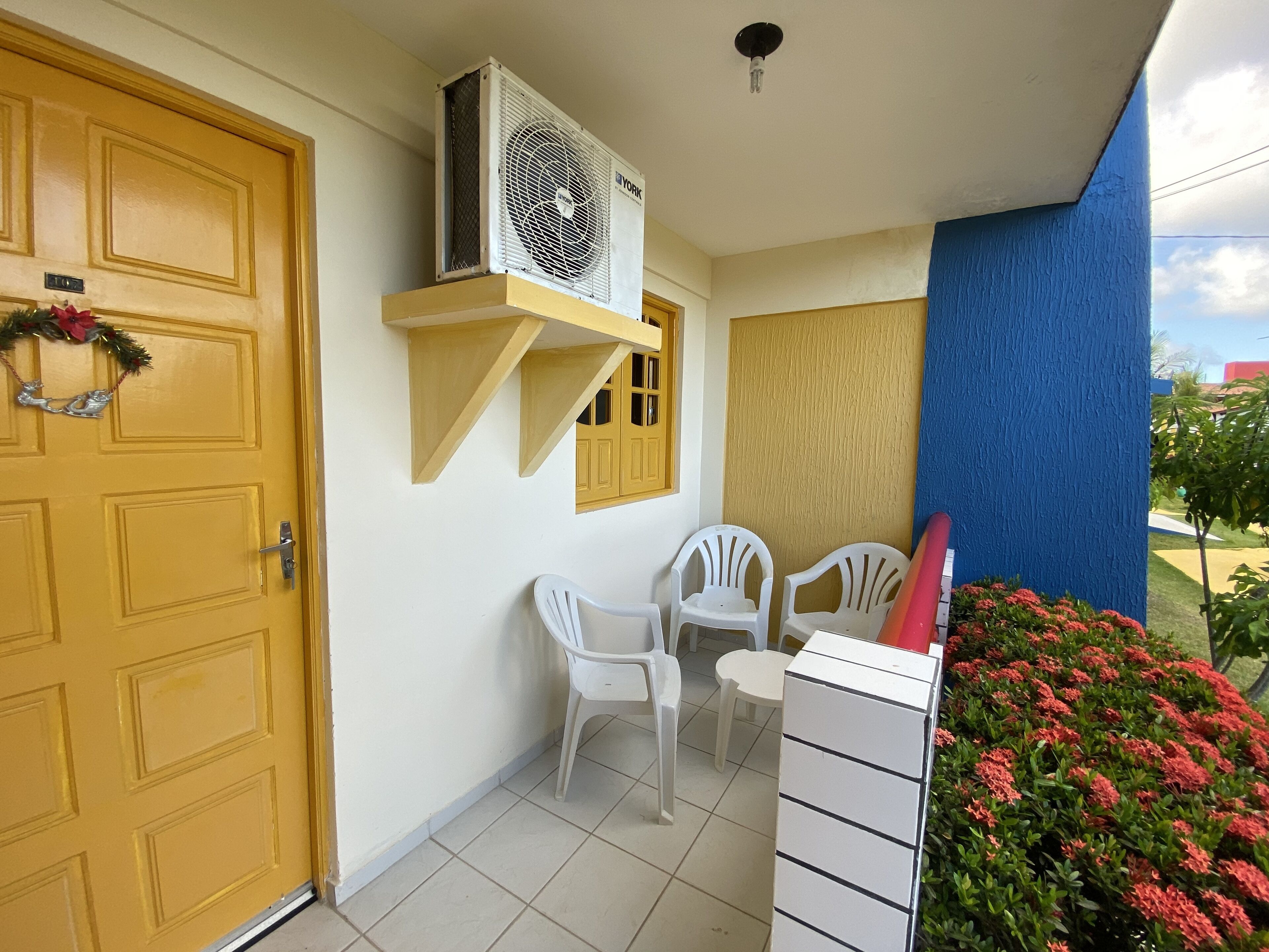 Een kamer met een gele deur, een witte airconditioning, twee witte plastic stoelen en een klein wit tafeltje.