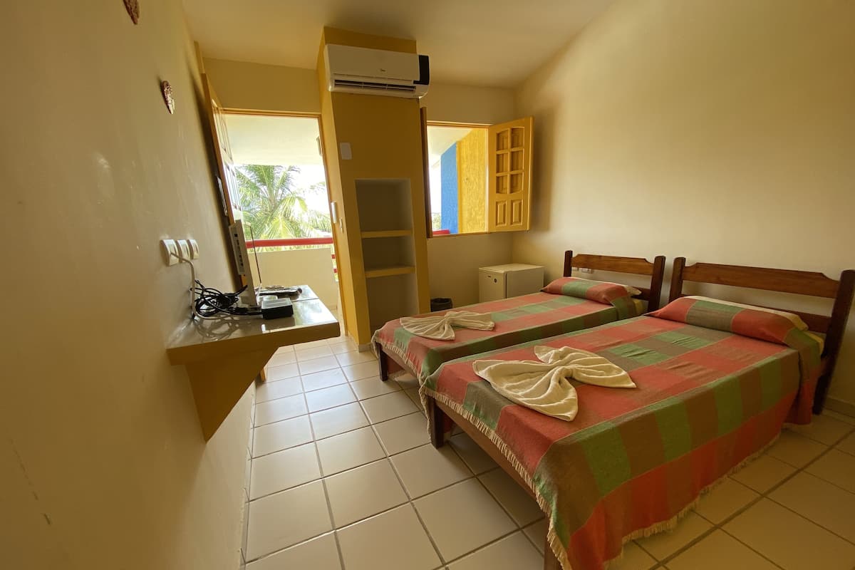 A room with two beds, a small desk, an air conditioner, and a window with a view of palm trees.