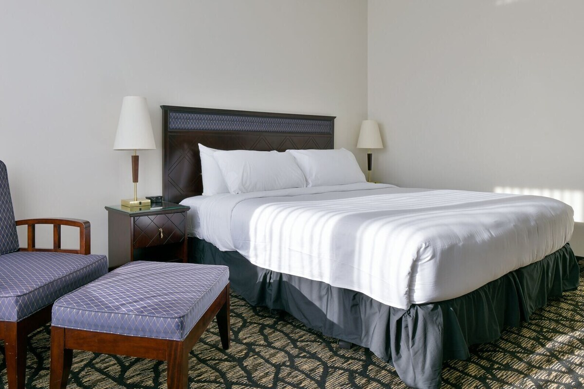 A hotel room with a bed, two bedside lamps, a chair, a small table, and a patterned carpet.