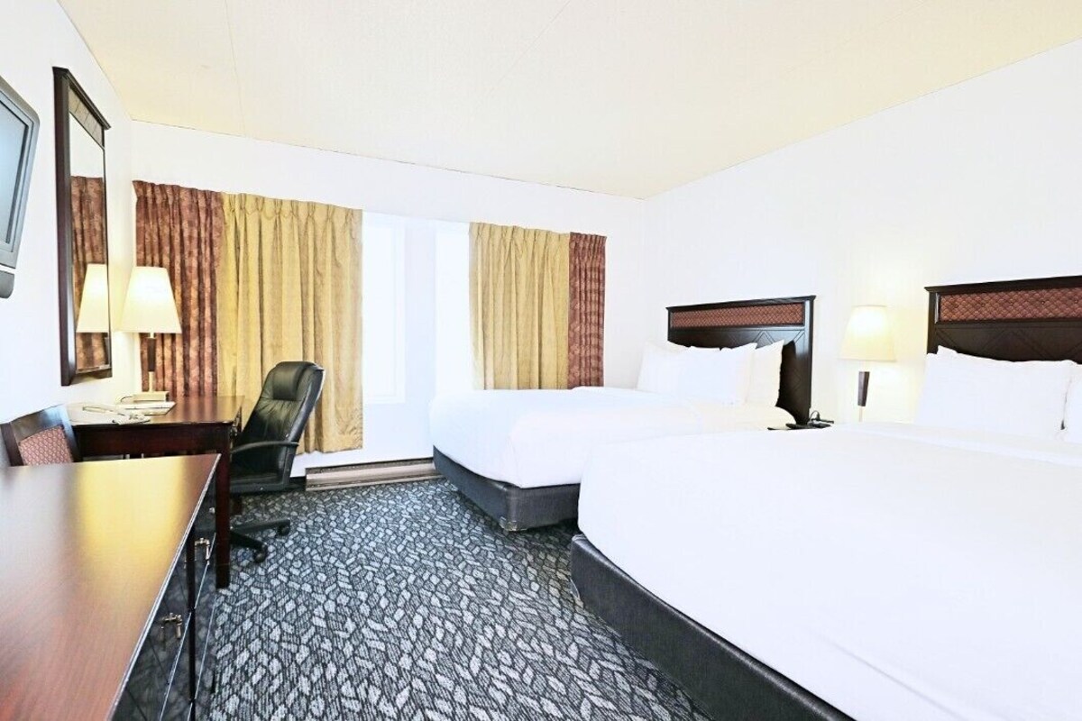 A hotel room with a desk, two beds, a chair, and a large window with curtains.